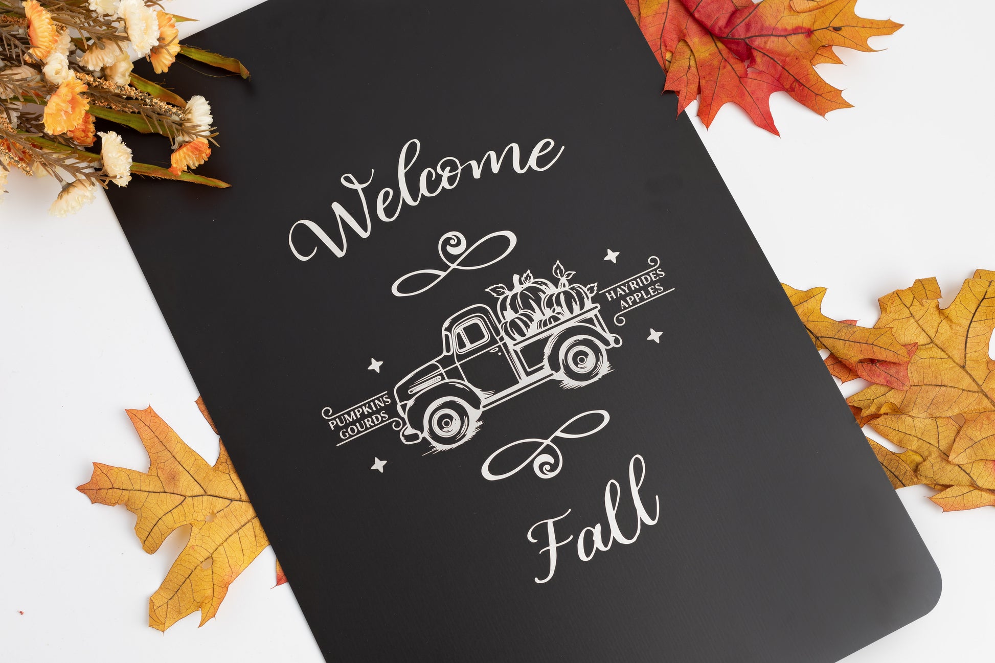 Black aluminum garden sign with 'Welcome Fall' text and vintage truck design, surrounded by autumn leaves.