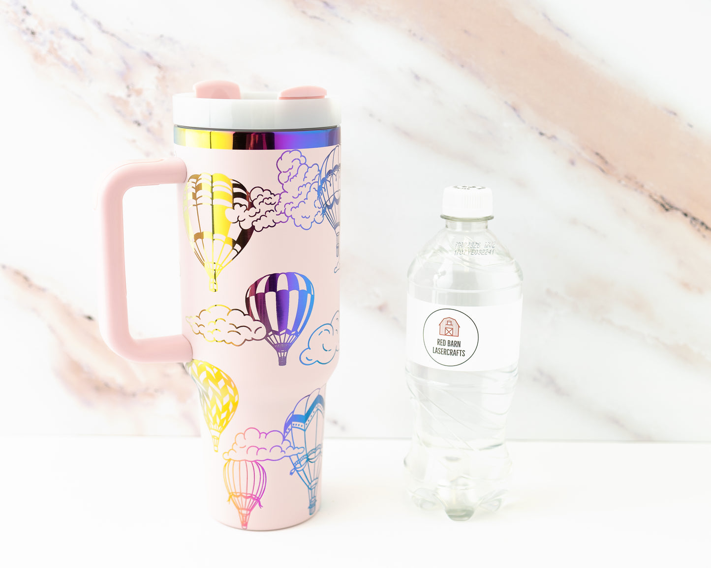 Laser engraved pink and rainbow 40oz stainless steel tumbler with hot air balloon and cloud design. Shown with a water bottle for size comparison.