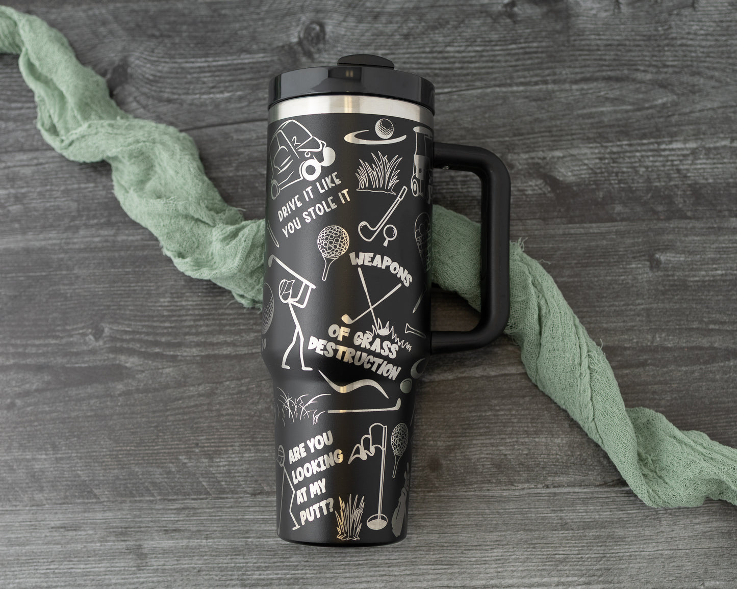 40 oz tumbler in black with golf funny sarcastic sayings and cartoon doodles wrapped around the tumbler. Flat lay on a grey background with a cloth.