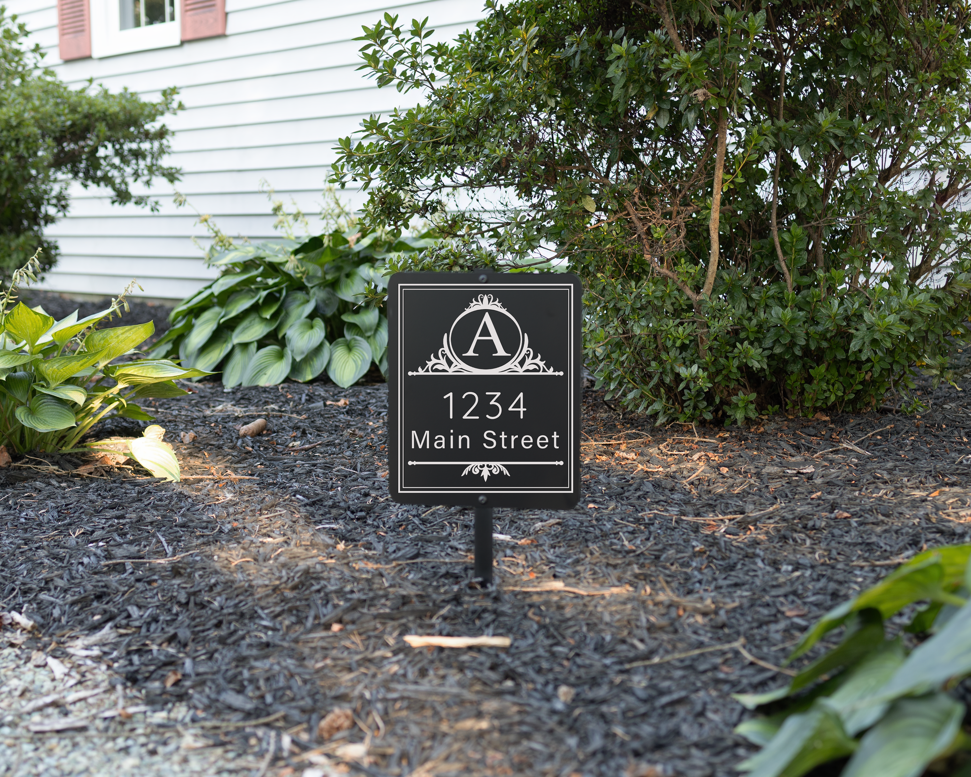 Black garden sign engraved with a decorative monogram and an address. Shown in a mulched flower bed.
